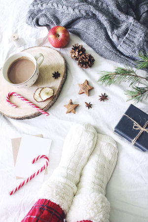 Christmas bed still life. Female legs in checkered pyjamas and woolen socks. Cup of coffee, candy canes and gift box. White linen bed sheet background with sweater. Winter flatlay, top view.の写真素材