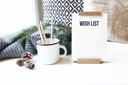Winter desktop still life scene. Wish list card, board and wooden pencils in mug standing near window on white table background. Christmas concept. Pine cones and fir branch on wool plaid.の写真素材