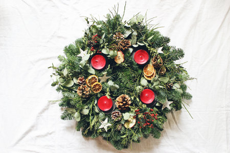 Christmas advent wreath isolated on white table background. Decorated by evergreen fir tree branches, eucalyptus leaves, wooden stars, pine cones, berries and red candles. Flat lay, top view.の写真素材