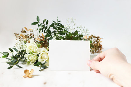 Bright feminine wedding stationery mockup scene. Closeup of woman hand holding paper place card. Bouquet of eucalyptus, dry hydrangea, white roses and gypsophila flowers on marble table background.の写真素材