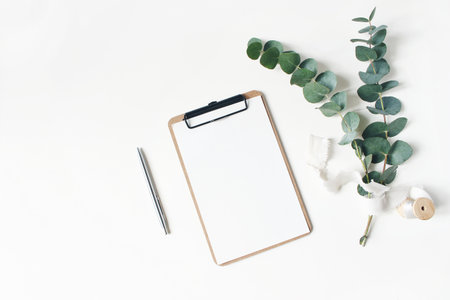 Feminine stationery mockup scene. Eucalyptus leaves and branches, silk ribbon, silver pen and blank greeting card in binder clip isolated on white table background. Flat lay, top view.の写真素材