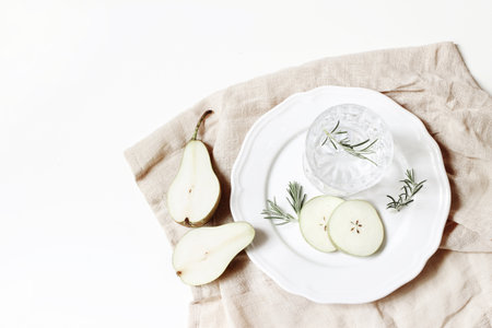 Pear cocktail, alcohol drink in a glasses with soda, fruit slices, and rosemary herbs leaves, porcelain plate, linen napkin on white table background. Summer refreshment concept. Flat lay, top view.の写真素材