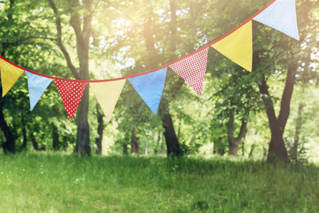 Colorful bunting flags hanging in park. Summer garden party. Outdoor birthday, wedding decoration. Midsummer, festa junina concept. Selective focus. Blurred grass and trees. Sunset haze.の写真素材