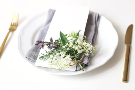 Close up of festive table summer setting.Golden cutlery, porcelain plate, blank menu card, napkin and herbs bouquet. Mediterranean wedding or restaurant concept. Selective focus, blurred background.の写真素材