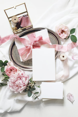 Feminine vertical wedding, birthday mock-up scene. Blank paper greeting cards, eucalyptus, pink roses, peony flowers on white table background.Golden glass box with silk ribbons. Flat lay, top viewの写真素材