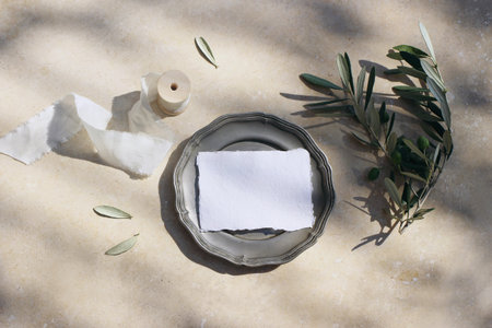 Summer wedding stationery mock-up scene. Blank cotton place card on vintage silver plate with olive branch, leaves and silk ribbon. Marble background in sunlight, shadows. Flat lay, top view.の写真素材