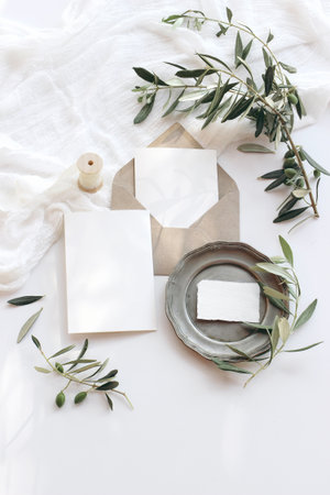 Summer wedding stationery mock-up scene. Blank greeting cards, envelope, vintage silver plate, olive branches and ribbon. White background with cotton table runner in sunlight. Flat lay, top view.の写真素材