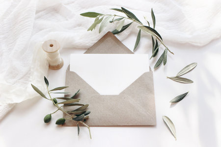 Feminine wedding stationery, desktop mock-up scene. Blank horizontal greeting card with craft envelope, silk ribbon and olive branches.White table background. Summer flat lay, top view.の写真素材