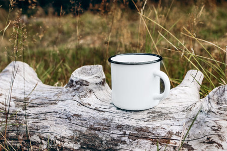 White metal mug sitting on old wood log. Blurred grass, meadow background. Outdoor tea, coffee time. Mockup of enamel cup. Lifestyle relax, trekking and camping concept.の写真素材