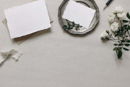 Stationery wedding table mockup scene. Moody composition with fading white rose flowers, silver...