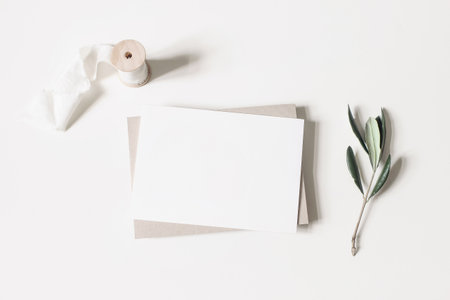 Summer wedding stationery mock-up scene. Blank horizontal greeting card, envelope, ribbon and olive branches isolated on white table background. Feminine Mediterranean flat lay, top view.の写真素材