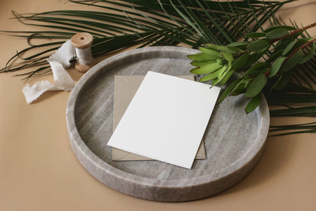 Summer tropical wedding stationery mock-up scene. Blank greeting card, envelope, ribbon and green palm leaves and leucadendron flowers on marble tray. Beige table background. High angle top view.の写真素材