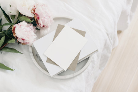 Wedding still life scene. Greeting card mockup scene, craft paper envelope, book on marble tray. Pink peony flowers on white linen table cloth. Vintage feminine styled photo, wooden floor. Flat lay.の写真素材
