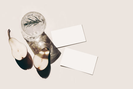 Summer stationery still life scene. Cut pear fruit, old cocktail glass, alcohol drink with ice cubes, rosemary herbs leaves isolated on beige table background. Business cards mockups. Flat lay, topの写真素材