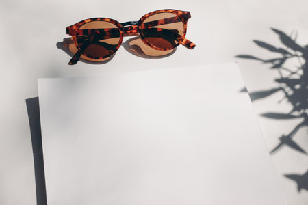 Summer stationery still life scene. Closeup of sunglasses with olive tree branch shadow in sunlight. White table. Blank paper card, invitation mockup scene. High angle view, no people.の写真素材