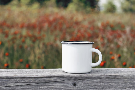 Close up of metal mug on old wooden table, board with defocused blooming mountain meadow. Outdoor tea, coffee time. Mockup of white enamel cup. Lifestyle relax, trekking and camping concept.の写真素材