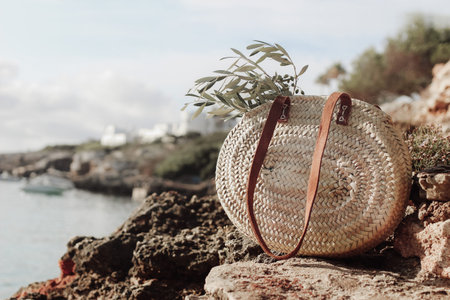 Summer vacation still life. French straw basket with olive tree branches on beach rocks. Blurred background with sea, white houses and boat. Selective focus. Mediterranean Mallorca island travel.の写真素材