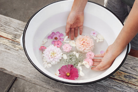 Child hands in white enamel bowl with garden flowers on old wooden bench. Pink roses, dahlia and cosmos floating in water. Spa, relaxation concept. Feminine summer tranquile lifestyle composition.の写真素材