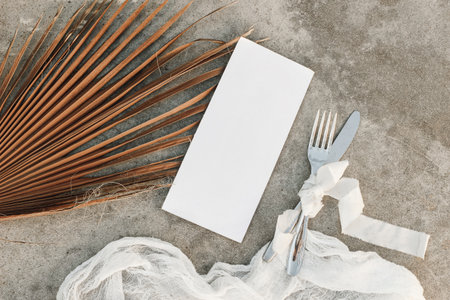 Tropical wedding ry still life. Closeup of blank menu card mock-up. Dry palm leaf, silver cutlery and muslin runner on grunge beige concrete background. Moody boho design. Flat lay, top view.の写真素材