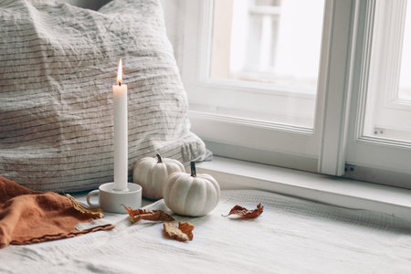 Autumn still life. Burning candle in white ceramic candleholder. Dry beech leaves and linen pillow near window. Moody composition with white pumpkins. Scandinavian interior.Thanksgiving, fall conceptの写真素材