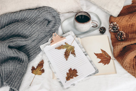 Fall breakfast still life. Cup of coffee, open book and notepad with colorful maple and oak leaves. White linen background. Linen pillows and grey sweater in bed. Autumn lifestyle concept. Top view.の写真素材