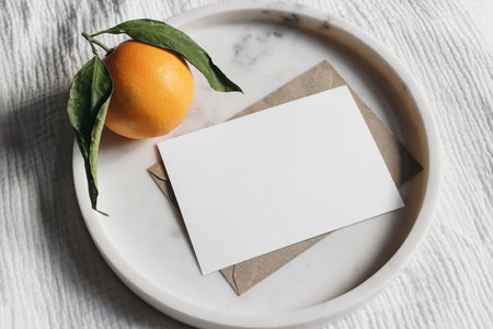 Summer wedding stationery mock-up scene. Blank greeting card, invitation. Craft envelope and orange fruit on elegant marble tray. White muslin tablecloth background. Top view.の写真素材