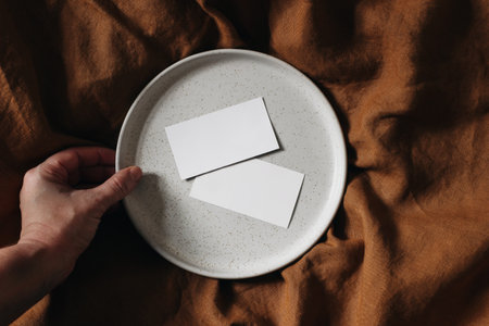 Two blank business cards mock-up scene. Female hand holding white ceramic plate. Wrinkled usty linen table cloth background. Advertisement, branding concept. Flat lay, top view.の写真素材