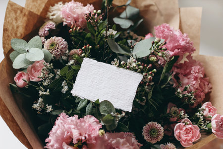 Moody feminine wedding, birthday mockup scene. Closeup of blank cotton paper card, invitation on pink floral bouquet. Eustoma, santini mums and carnations with ucalyptus. Selective focus, top view.の写真素材