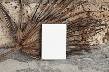 Summer wedding stationery, birthday mock-up. Empty greeting card agaist stone wall. Dry palm tree leaf. Grunge concrete background in sunlight. Front view. Tropical vacation concept.の写真素材