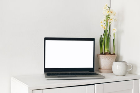 Spring home office still life composition. Laptop mockup, blank computer screen. Yellow daffodils in flower pot. Cup of coffee on wooden cupboard, table. White wall background. Scandinavian interior.の写真素材
