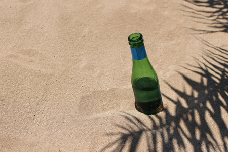 Green empty glass bottle of water in golden sand with palm leaf shadow. Summer vacation. Refreshment idea. Trash on the beach. Concept of garbage seashore nad ocean pollution.の写真素材