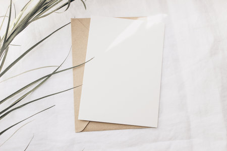Modern summer stationery still life. Dry palm leaf on white linen table cloth. Blank greeting card mock up scene with craft envelope in sunlight. Flat lay, top view. Tropical design. Sun flare leaks.の写真素材