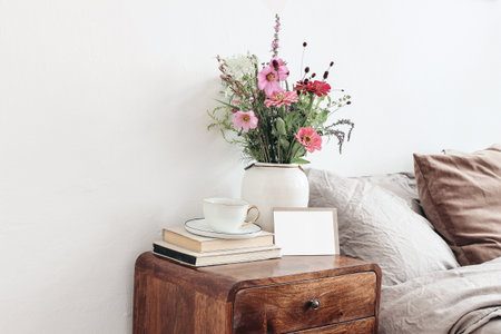 Cup of coffee and books on retro wooden bedside table. Blank greeting card mockup. White ceramic vase. Buquet of cosmos and zinnia flowers. Beige linen, velvet pillows in bed. Scandinavian interior.の写真素材