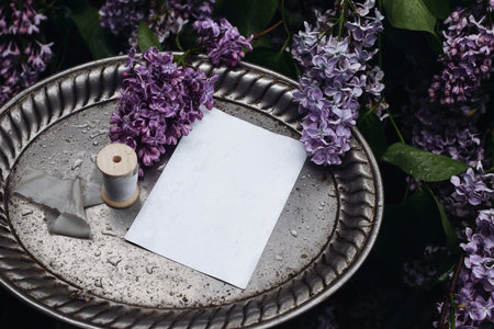 Moody spring wedding stationery mockup scene. Blank greeting card, invitation on vintage silver tray. Purple lilacs flowers and rain drops. Wet silk ribbon. Blooming Syringa bush in the garden. Top.の写真素材