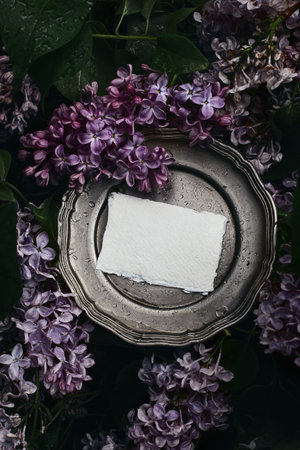 Moody spring wedding stationery mockup scene. Blank greeting card, invitation on vintage silver plate. Purple lilacs flowers and rain drops, blooming Syringa bush in the garden. Vertical flat lay, top viewの写真素材