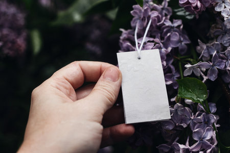 Womans hand holding blank gift tag, label. Purple lilacs blossoms in the garden. Moody spring mockup. Birthday celeberation. Business, sale concept. Dark floral still life, blurred background.の写真素材