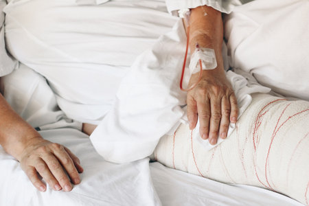 Injured old woman lying in bed with white blanket in hospital. Closeup of hands with vein cannula and bandaged leg. Emergency surgical treatment. Ilness, healing and recovery concept, elderly care.の写真素材