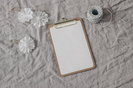 Winter festive still life. Christmas ornaments, decoration. White paper snowflakes and decorative gift rope on beige linen tablecloth. Blank paper sheet mockup. Wooden clipboard. Flat lay, top view.の写真素材