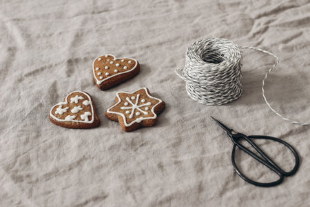 Christmas festive still life. Gingerbread star, heart cookies with sugar icing and decorative gift rope. Beige linen tablecloth. Gift wrapping. Old black scissors. Selective focus, blurred background.の写真素材