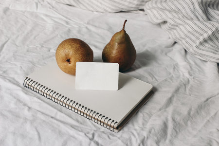 Couple of yellow pear fruit on blank notebook, diary. Business card mockup. Autumn, summer breakfast in bed composition.White striped linen bedding. Feminine lifestyle composition. Rustic interior.の写真素材