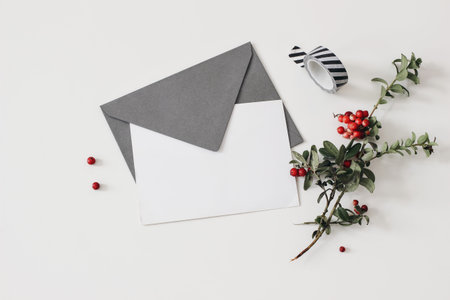 Blank greeting card mockup with craft envelope and washi tape. Christmas stationery, wish list. Red lingonberries fruit with green leaves isolated on white table background. Winter flat lay, top viewの写真素材