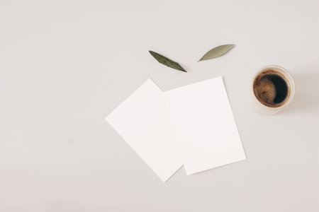 Blank greeting cards and olive tree leaves on beige background. Wedding invitations mockups scene. Cup of coffee. Breakfast concept. Flat lay, top overhead view. Working table with office equipment.の写真素材