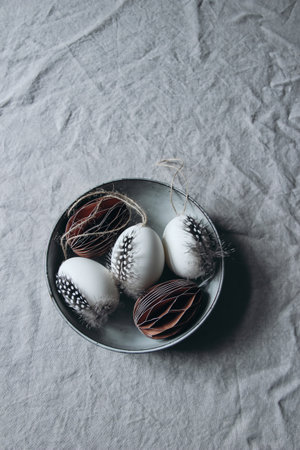Moody spring still life scene. Easter composition. Bowl of folded paper ornaments and hen eggs decorated by feather of guinea fowl bird. Gray linen table cloth background. Neutral vertical flat lay. Top view.の写真素材