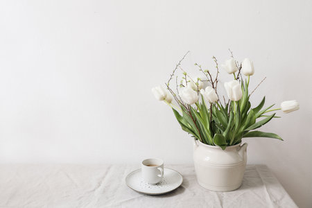 Feminine Easter, spring still life scene. Cup of coffee and floral bouquet. White ceramic vase on table. White tulips flowers, cherry tree branches on linen tablecloth. Styled interior. Copy space.の写真素材