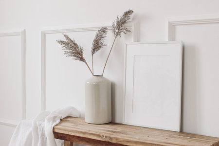 Elegant home interior decor still life photo. Vase with dry reed, grass on old wooden bench. Blank white picture frame mockup. Wall moulding background, trim decor. Empty copy space. Side view.の写真素材