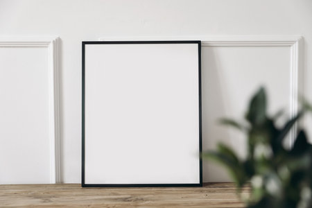 Blank black picture frame mockup on old wooden bench, table. Wall moulding background, trim decor. Elegant home interior decor, still life photo. Art dispaly. Blurred green leaves, plant foreground.の写真素材