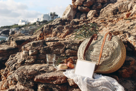 Summer vacation. Picnic breakfast on beach. Straw basket, olive tree branches. Blank mockup card. Glass of water, peach fruit. Blurred background with sea, white houses. Selective focus. Mallorca.の写真素材