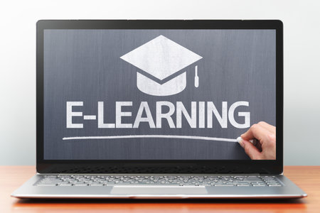 E-learning concept. Laptop computer on wooden desk. Teacher writing on blackboard. Front view image.の写真素材