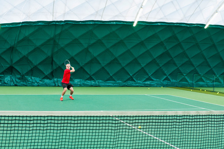 Closeup of professional tennis player dressed in red t-shirt, black shorts and red sport shoes. Man holds tenis racket and playing tennis on court indoor.の写真素材