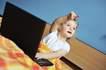 cheerful little girl with laptop sits on bed of parentsの写真素材